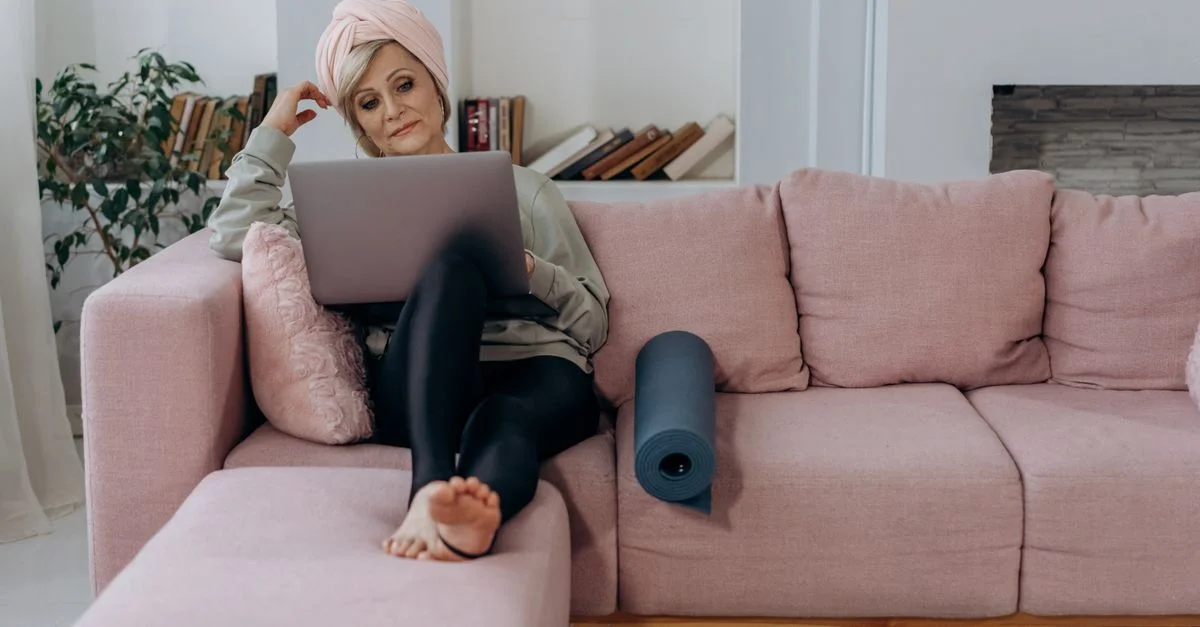 Femme senior assise sur un canapé rose utilisant un ordinateur portable chez elle, avec un tapis de yoga et un turban.
